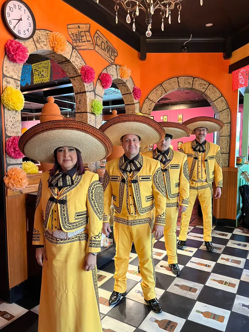 Cuatro integrantes del mariachi con traje amarillo bordado y sombrero posan en un restaurante decorado con colores y piso de cuadros