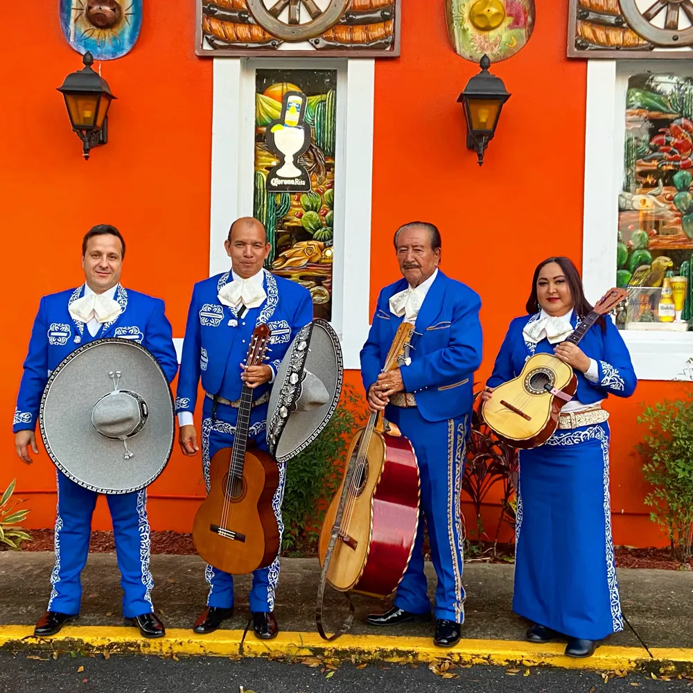 Cuatro integrantes del mariachi con traje azul bordado, con sus instrumentos y sombreros, frente a una pared naranja con faroles