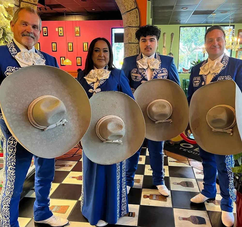 Cuatro integrantes del mariachi con traje azul y bordados claros sostienen grandes sombreros grises en un restaurante con piso de cuadros