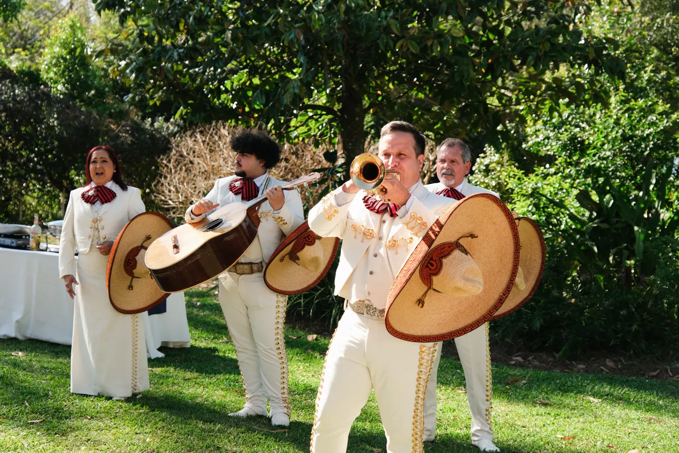 Músicos del mariachi con traje claro al aire libre: uno toca la trompeta en primer plano y otros sostienen guitarras y sombreros, con árboles y una mesa al fondo