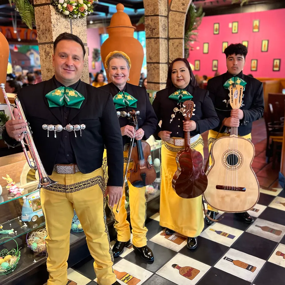 Cuatro integrantes del mariachi con chaqueta negra y pantalón amarillo, con trompeta, violín, vihuela y guitarrón, en un restaurante