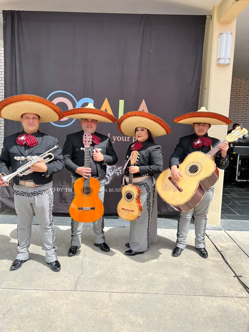 Cuatro integrantes del mariachi con traje gris y sombrero posan con trompeta y guitarras frente a un fondo oscuro donde se lee la palabra OCALA