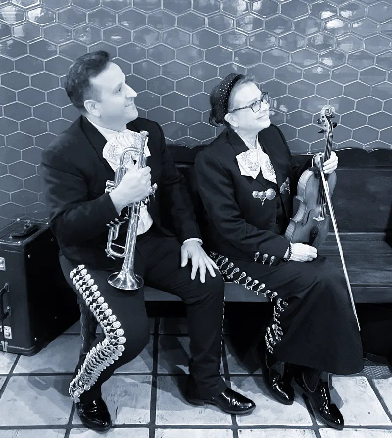 Fotografía en blanco y negro de un músico con una trompeta y una música con un violín, sentados en una banca frente a una pared de azulejos
