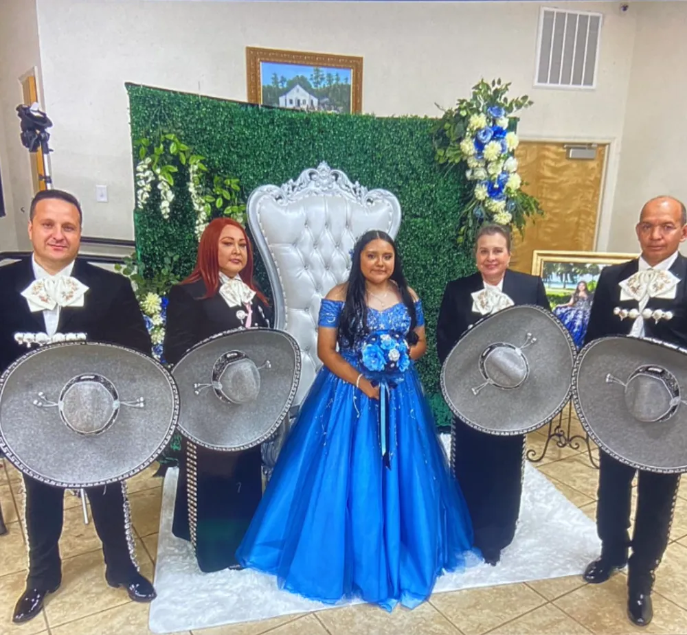 Quinceañera con vestido azul junto a cuatro integrantes del mariachi con traje de charro negro que sostienen sus sombreros plateados, en un salón decorado