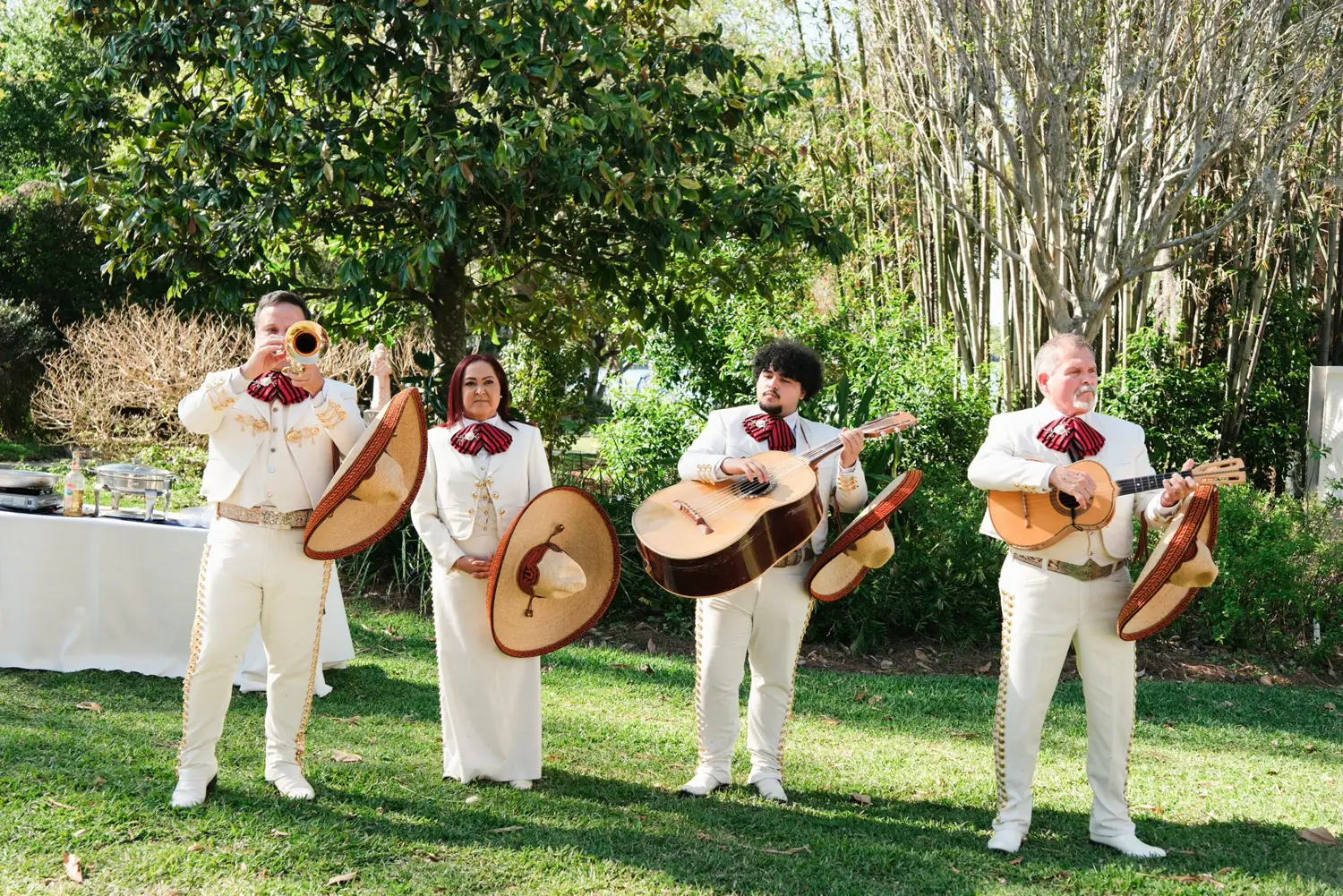 Cuatro músicos del mariachi con traje claro tocando guitarras y trompeta sobre el césped de un jardín, con árboles y mesas con mantel blanco al fondo